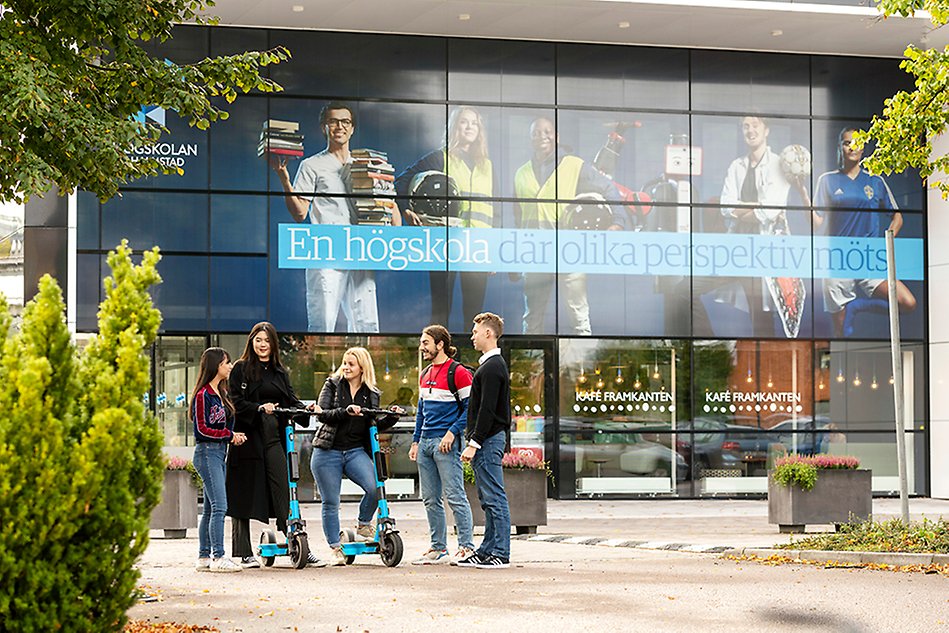 En grupp unga människor står utanför Högskolans entré. Två av dem står på en elsparkcykel. Foto.