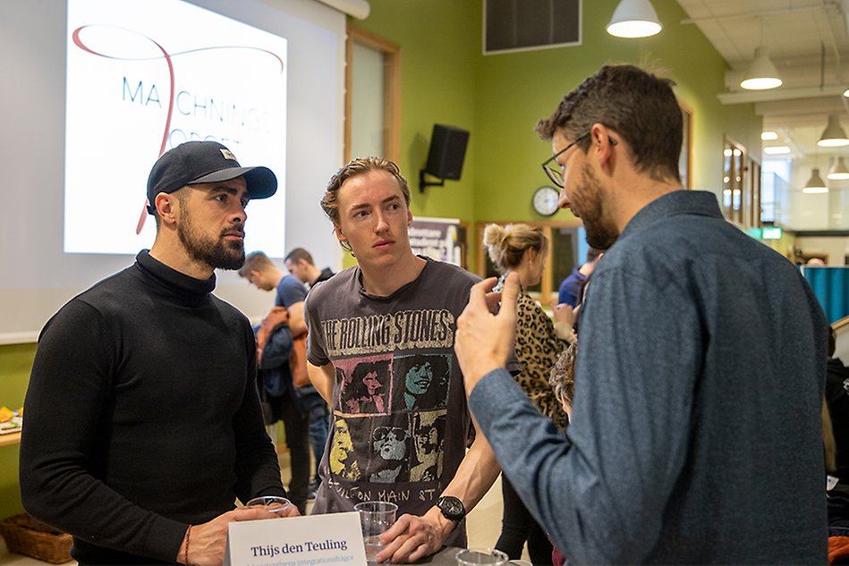 Två manliga studenter står och lyssnar på en man vid ett runt bord