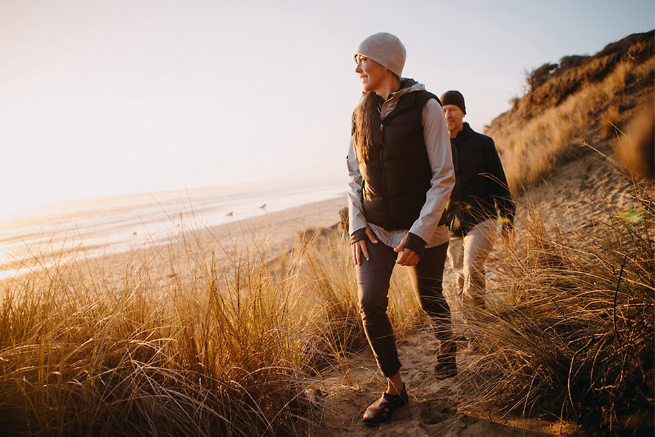 Two people dressed in winter clothes walk on a beach, the light from the sunset reflects on them from the left. Photo.