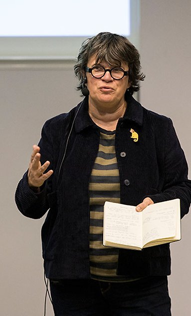 En kvinna talar från scenen i hörsalen på högskolan. I sin ena hand håller hon ett anteckningsblock där handskrivna anteckningar syns.