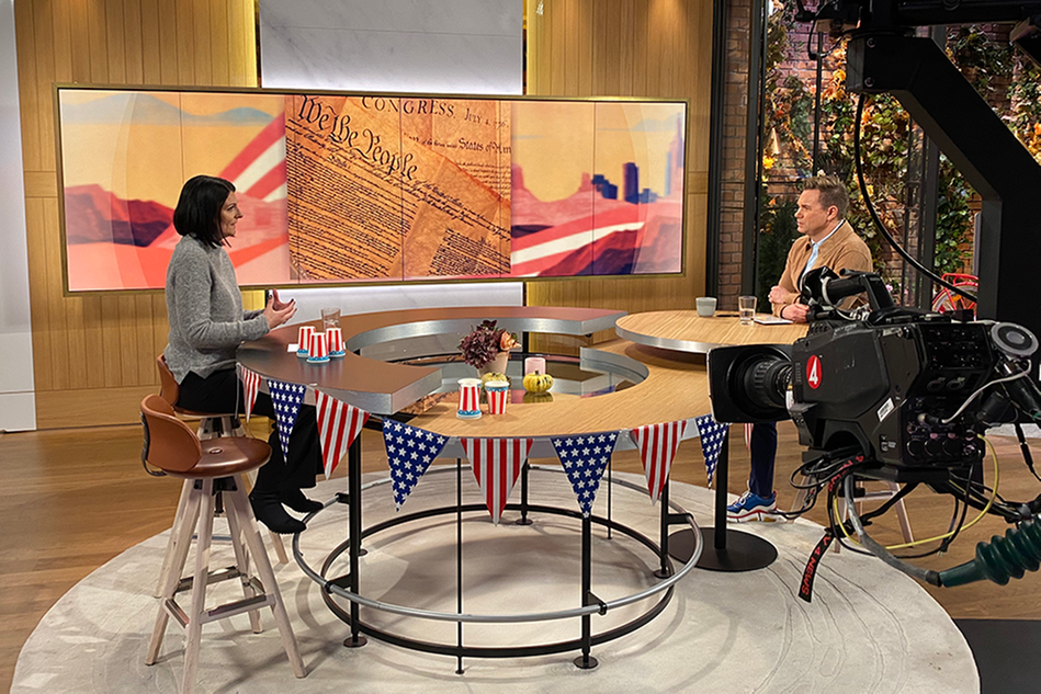 A TV studio with a man and a woman talking to each other. A large TV camera in the foreground. Photo