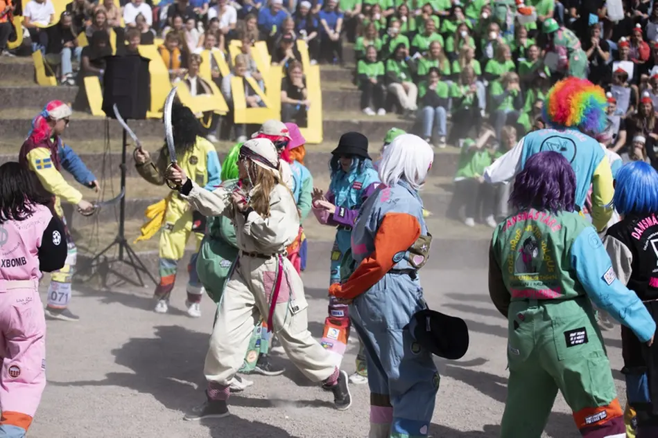 Ett flertal människor i masker och färgglada overaller dansar inne på en amfiteater utomhus. Foto.
