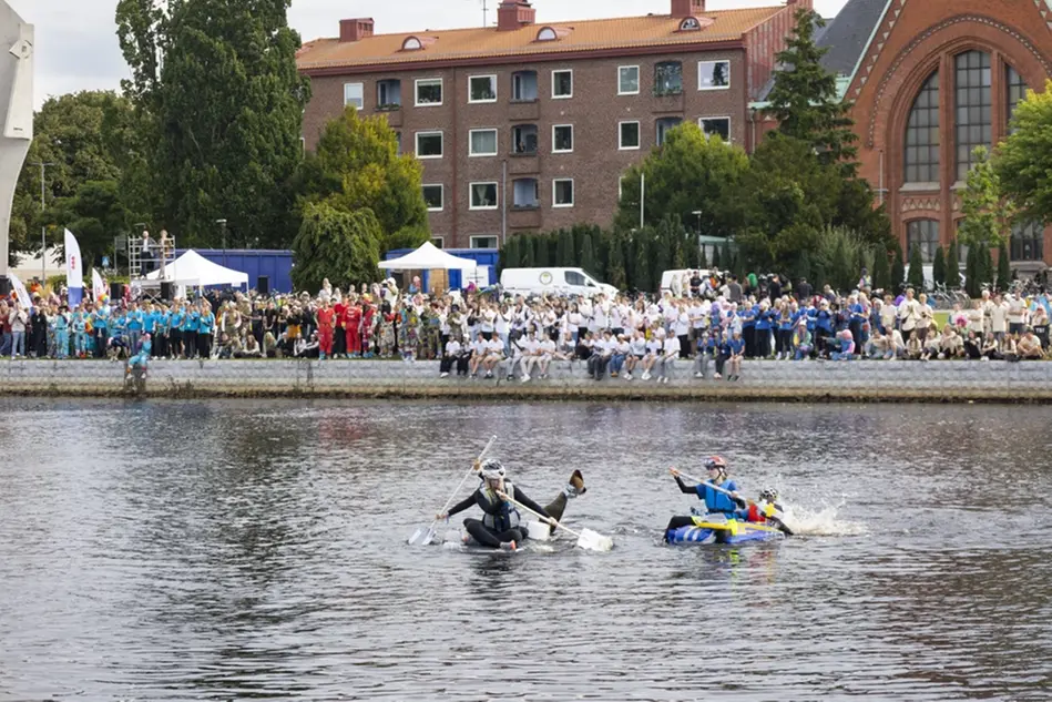 Fyra unga människor paddlar över en flod i två båtar. På flodkanten står en stor publik och tittar på. Foto.