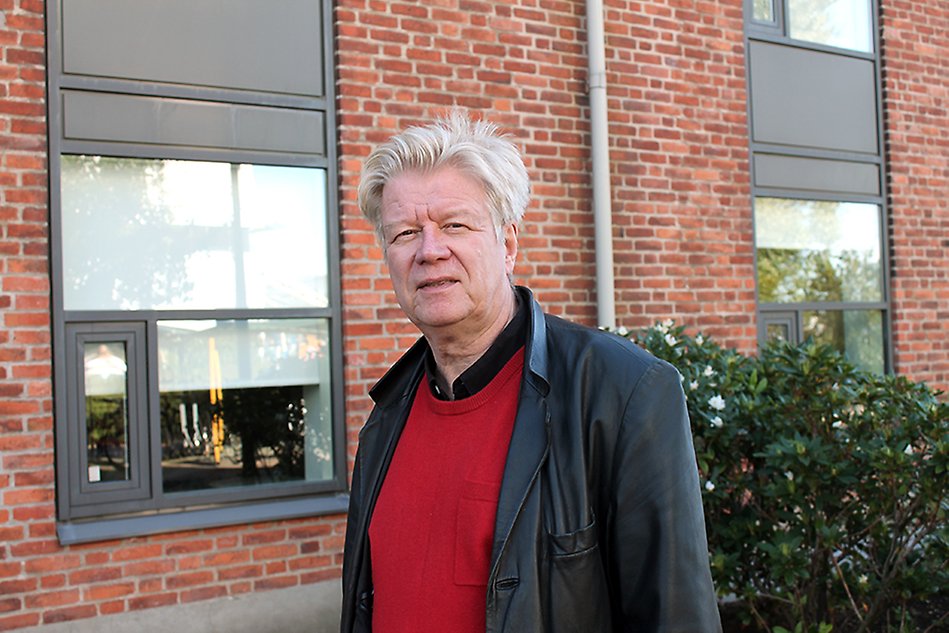 A man wearing a black jacket and red top standing in front of a red brick building with windows. Photo.