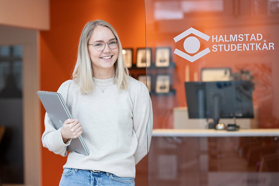 En leende ung kvinna lutar sig mot en glasvägg och håller en laptop i handen, i bakgrunden syns en orange vägg. Foto.