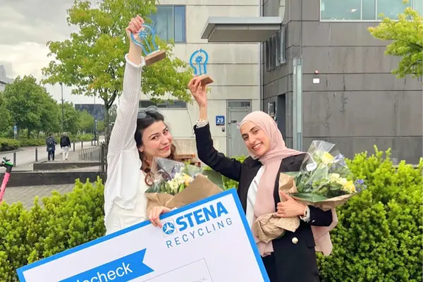 Två studenter ler mot kameran framför ett grönt buskage. De lyfter sina statyetter högt medan de håller i blombuketter och en värdecheck. Foto.