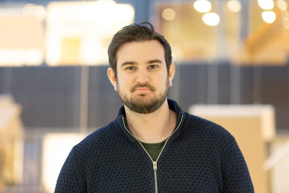 Mörkhårig student ler mot kameran. Han bär en grön t-shirt och mörkblå överdragströja. Foto.