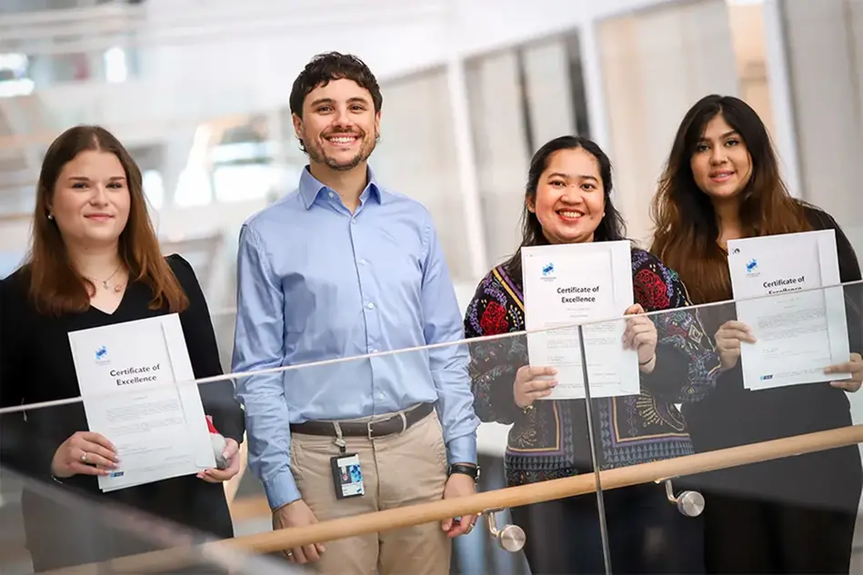 A group of people standing in a bright campus envionment, one of them holding a certificate. Photo.