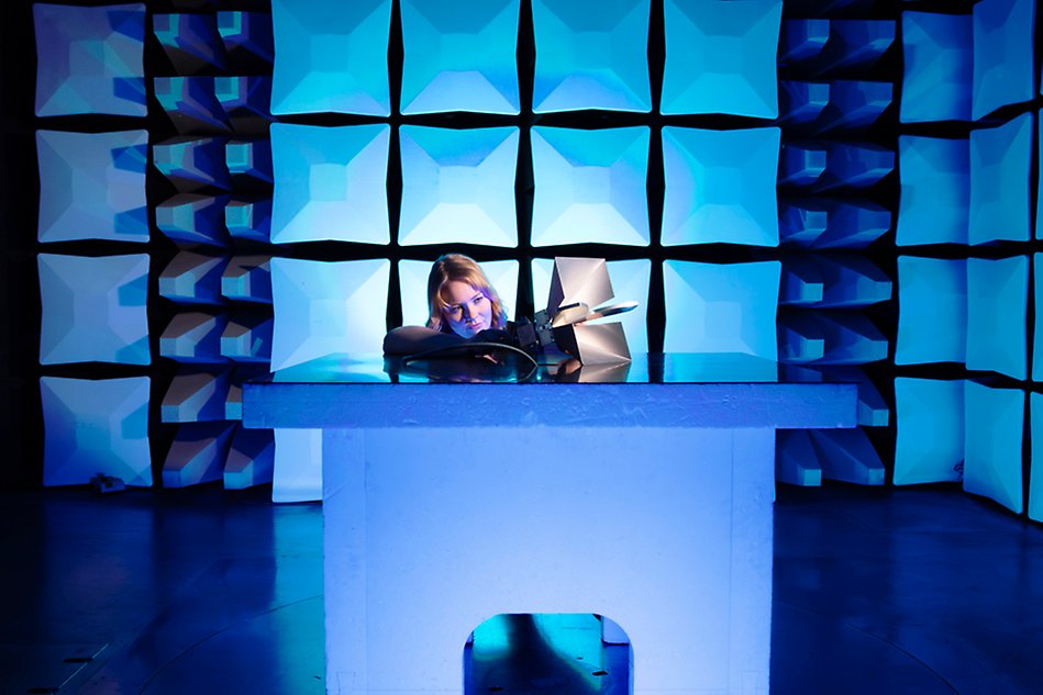 Kvinna står vid bord inne i ljudisolerande kammare och håller i en elektronisk apparat medan utanför sitter en man och studerar en datorskärm. Foto.