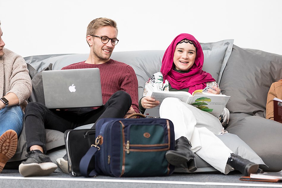 A younger man and a younger woman are seated in bean bags, each one of them with a computer in front of them. The man, who has glasses and light hair, looks at the woman's computer screen. The woman looks into the camera and is wearing a dark pink shawl around her head. Photo.