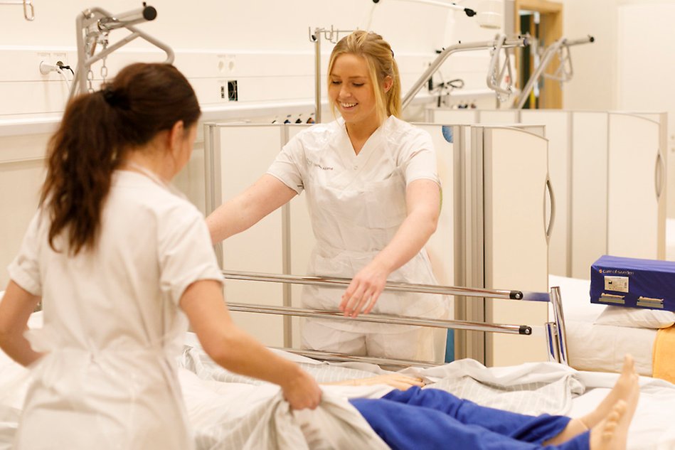 Two women in nurses' clothing are standing by a hospital bed, a dummy is lying in the bed
