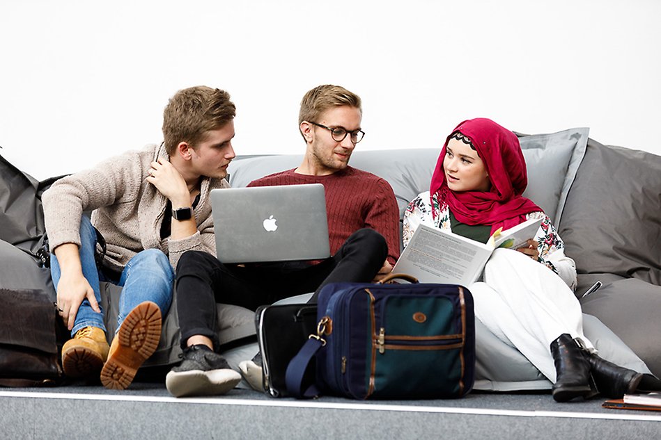 Tre studenter sitter på rad i bean bags på ett grått golv och diskuterar med varandra. Studenten i mitten har en dator i knäet och studenten till höger visar något i en bok för de andra, som tittar intresserat i boken. Foto. 