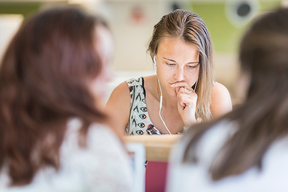 En student med ljust, mellanlångt hår sitter med handen framför munnen och ser fundersam ut. Hen har vita hörlurar på sig och tittar ned. I förgrunden syns två suddiga silhuetter. Foto.