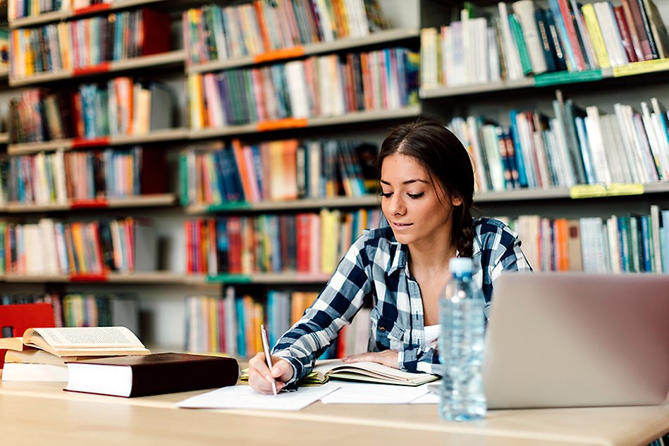 Ung kvinna sitter och skriver i ett block vid bord i ett bibliotek. Foto.