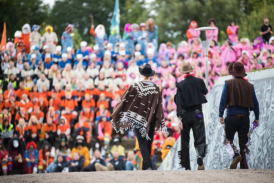 Tre män i cowboykläder går mot åskådarläktare fylld med nya studenter i färgglada t-shirtar. Foto.