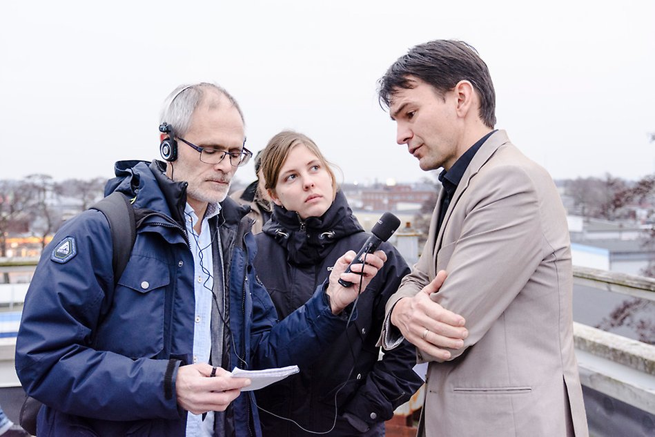 En man blir intervjuad av en man med mikrofon och hörlurar, en kvinna iakttar. Foto.