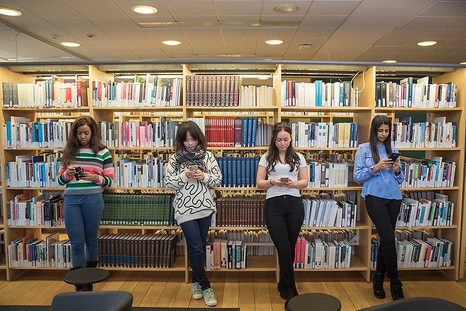 Fyra studenter står på rad framför en bokhylla full med böcker som de lutar sig mot. Alla studenter tittar ner i sin egen mobiltelefon. Foto. 