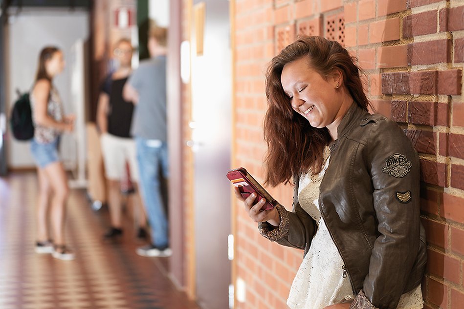 A woman leans against a red brick wall and looks down at her cellphone. In the background stands a group of other students. Photo. 