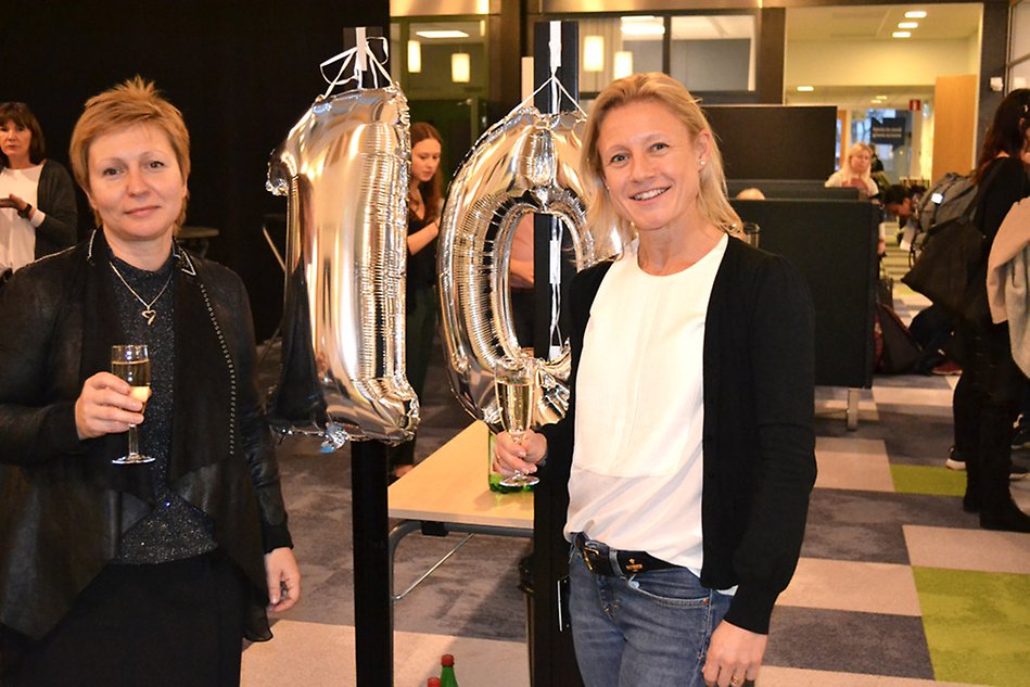 Två kvinnor står med bubbelglas framför silverballonger i form av talet 10.