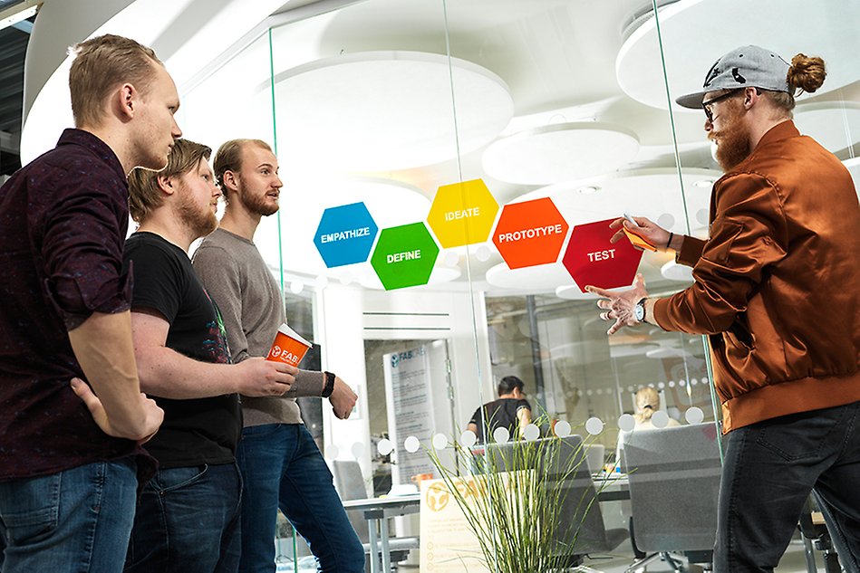 Man pointing at colored stickers on a wall, while three other men watch. Photo. 