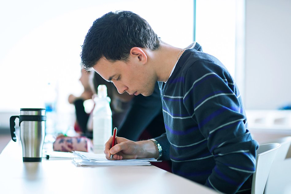 A man sitting down by a table, writing. Photo.