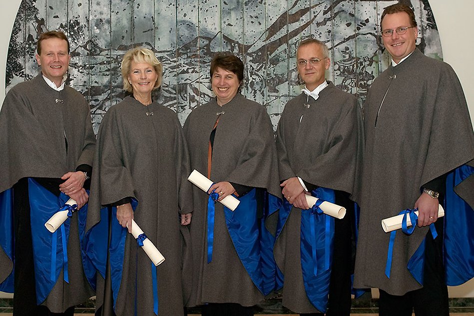 Fem personer i grå professorscaper framför mönstrad bakgrund. Foto.