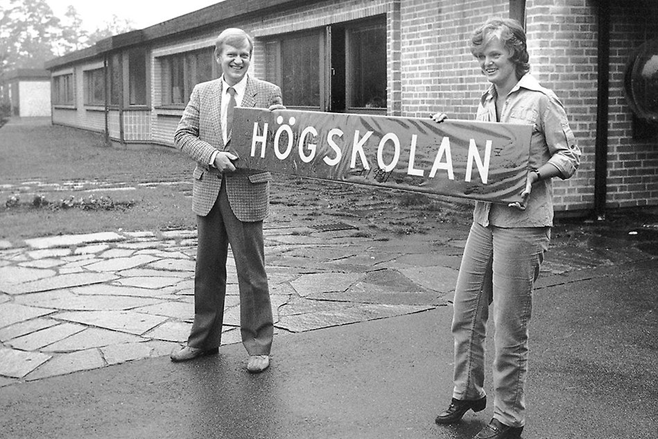 Two people standing outside building holding a sign. Black and white photo.