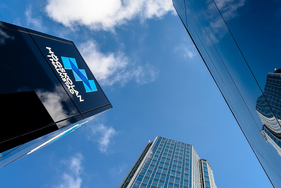 Blue sky with the top of black pillars with the University's logo and two buildings in the foreground. Photo.