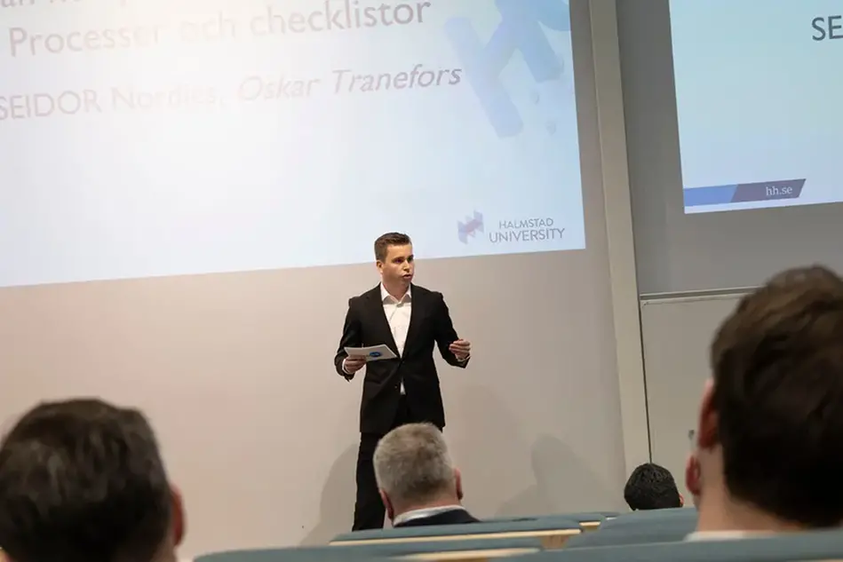 Person i kostym står med talkort och talar inför publik.