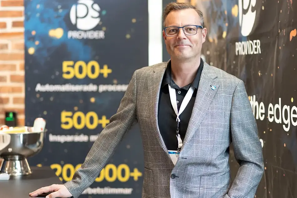 Man i glasögon, svart pikétröja och rutig kavaj lutar sig mot ett bord och ler mot kameran.