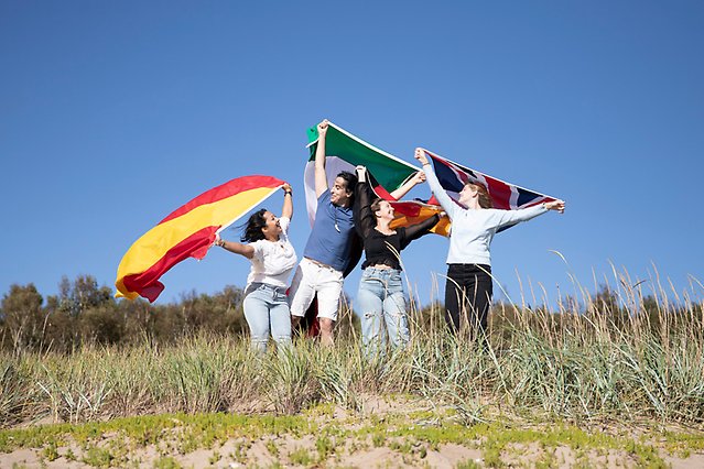 Fyra studenter håller i flaggor ovanför huvudet och ler mot varandra. De står på stranden. Foto.