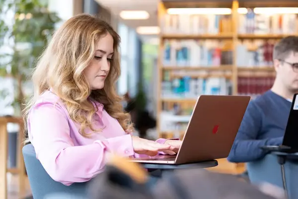 A student wearing a pink shirt sitting in a library setting with her laptop on her lap. She looks focused. Photo.