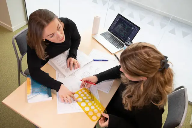 Två studenter diskuterar vid ett bord med studiematerial på. Foto.