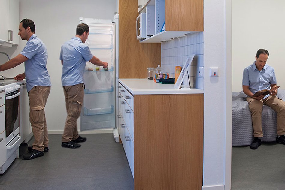 A view of the kitchen and bedroom of a flat, the same man is seen doing different things in the different rooms.