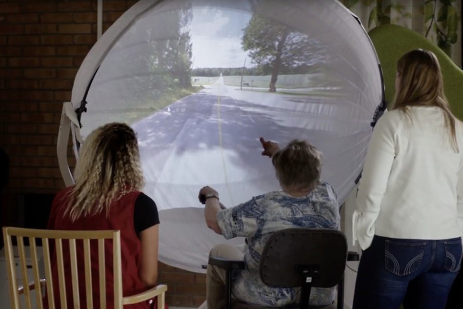 An elderly woman is sitting on an exercise bike in front of a curved screen showing a view of the outside, two other women are standing next to her.