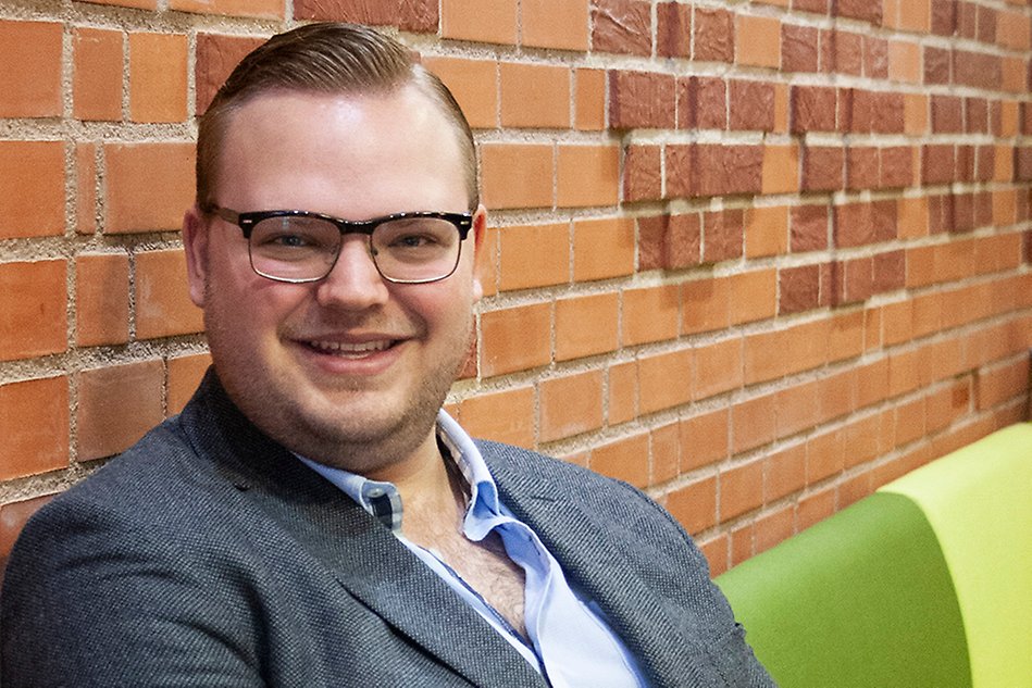Leende man med kort, ljust hår, glasögon och kavaj sitter framför en röd tegelvägg. Foto.