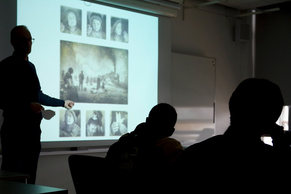 Man standing next to a projection showing old images of children and a train, an audience is facing the screen