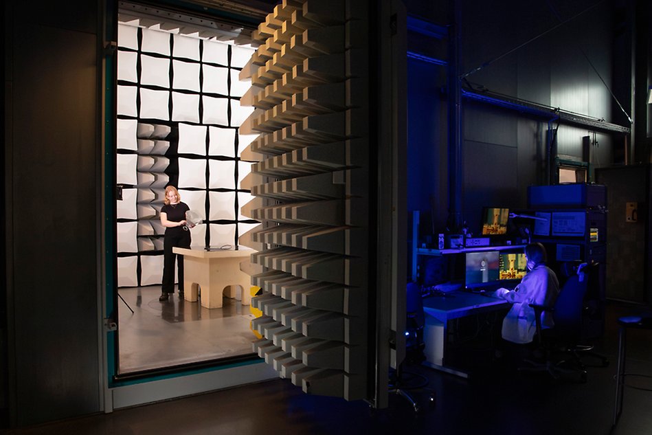 A person in a test chamber with an open door. On the right, a person can be seen at a computer outside the chamber. Photo.