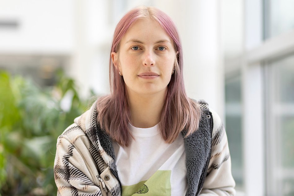 Kvinna sitter i korridor och tittar in i kameran, i bakgrunden ser man gröna växter. Foto.