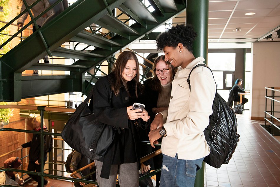 Tre personer står intill varandra och tittar ner i en mobil. I bakgrunden en trappa och trapphall. Foto