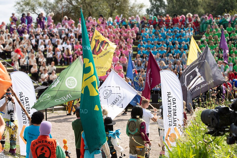  Flags in different colors are waived in the air inside a smaller round arena. In the background there's a crowd with colorful t-shirts sitting in stands. Photo