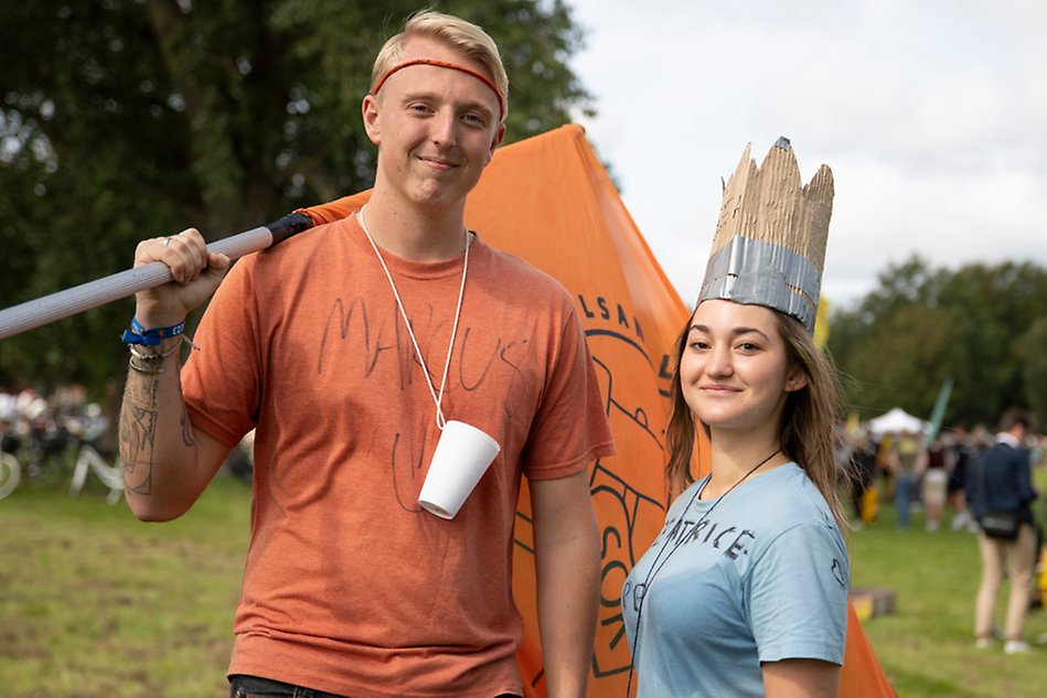 A young man with an orange t-shirt and a you woman with a light blue t-shirt are standing next to each other smiling. The man holds a flag and the woman has a hat made of paper. Photo.
