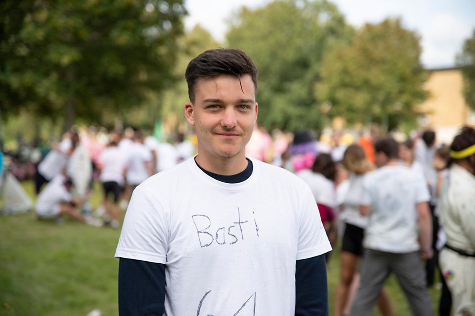 A young man with brown hair and brown eyes in a white t-shirt looks at the camera and smiles. Photo.