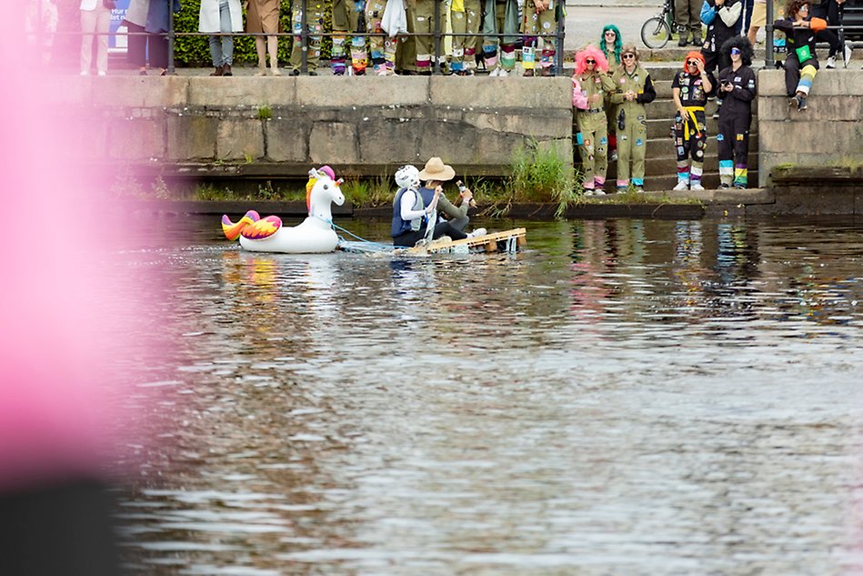 Två personer, en med hatt och en med hockeyhjälm, sitter på en träpall i en flod i stadsmiljö och paddlar. Foto.