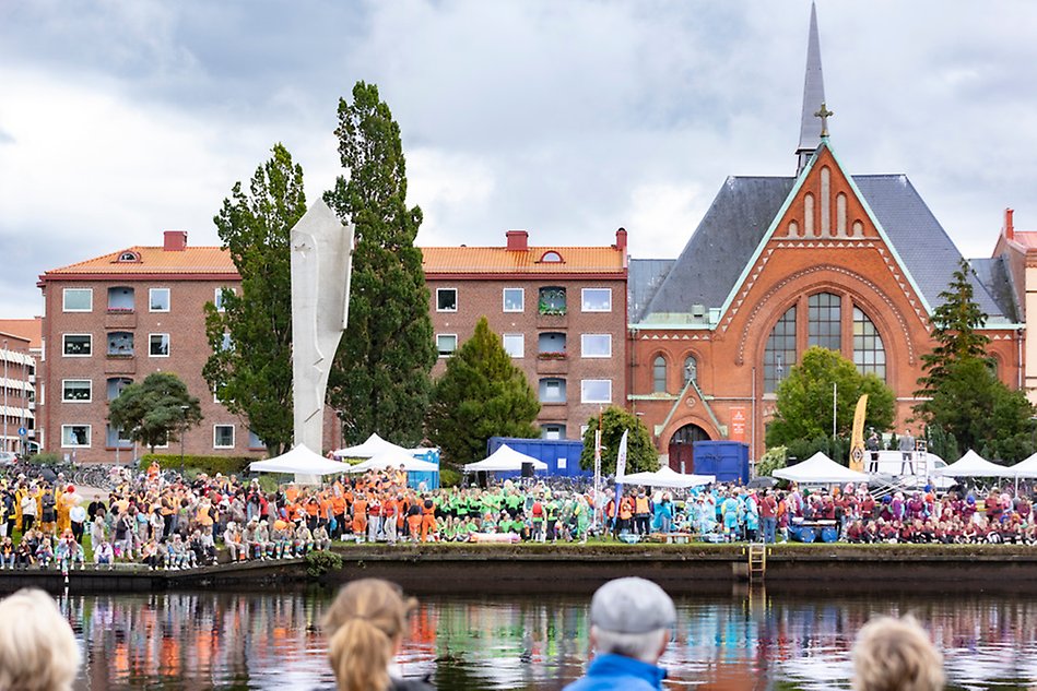 Utsikt över en flod i stadsmiljö. På andra sidan vattnet står en folkmassa samlad i en mindre, lummig park. Foto.