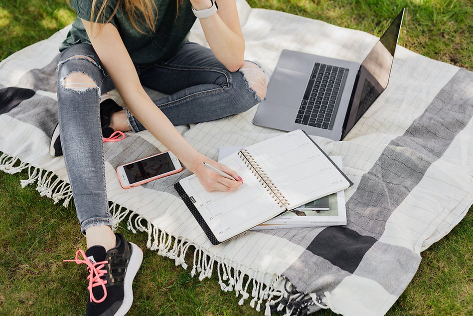 En person som sitter på en filt på gräset med en dator, en skrivbok och en mobil. Fotografi.