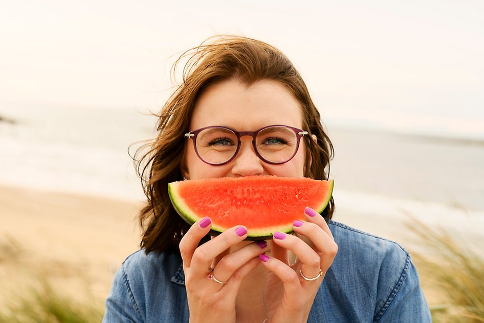 En person på en strand som håller upp en vattenmelonsklyfta framför ansiktet så att det ser ut som en glad mun. Fotografi.