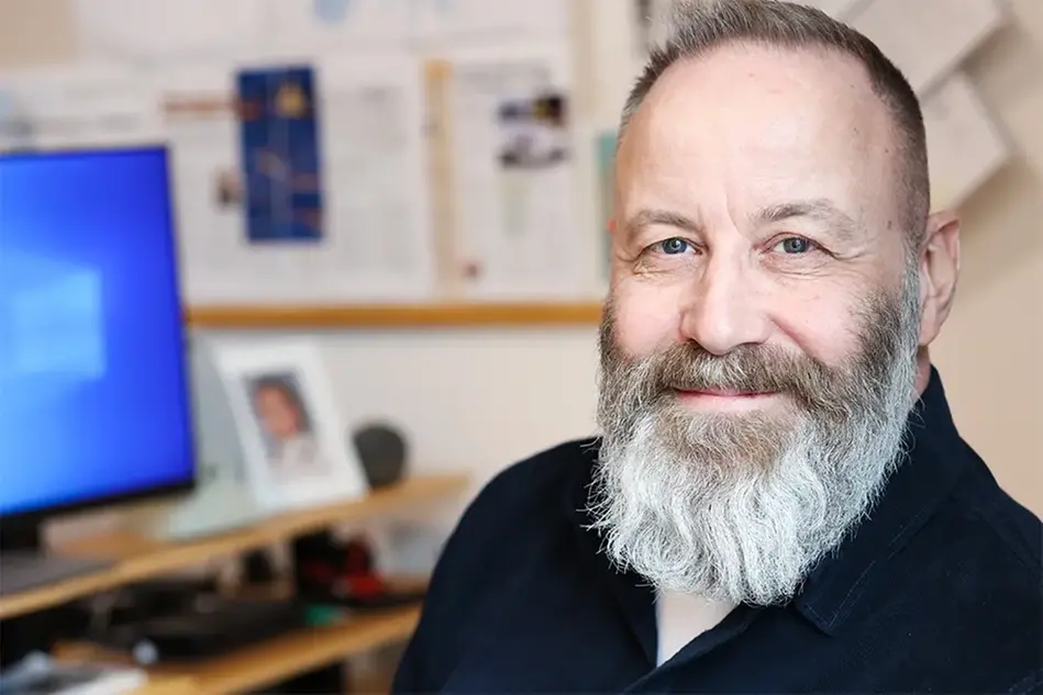 A bearded man sits by a computer and looks into the camera. Photo.