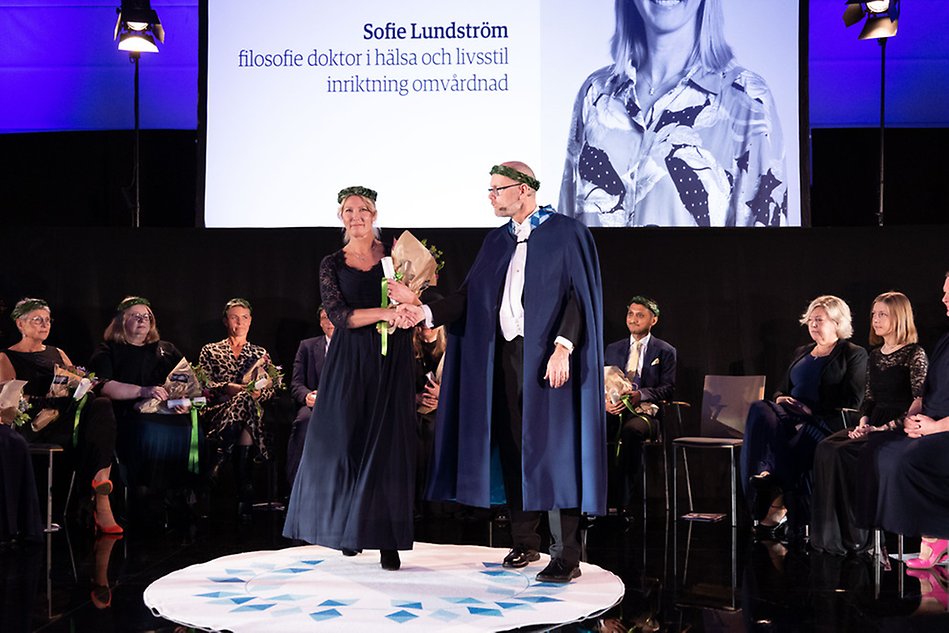 A woman wearing a long dress with a wreath on her head being led across a white, circular rug by a man wearing formal clothes and a cape. 