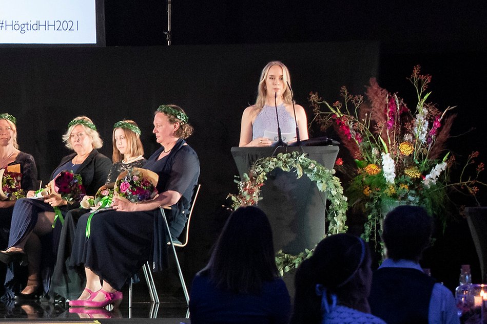 A woman on a podium surrounded by flowers, several other people sitting behind her.